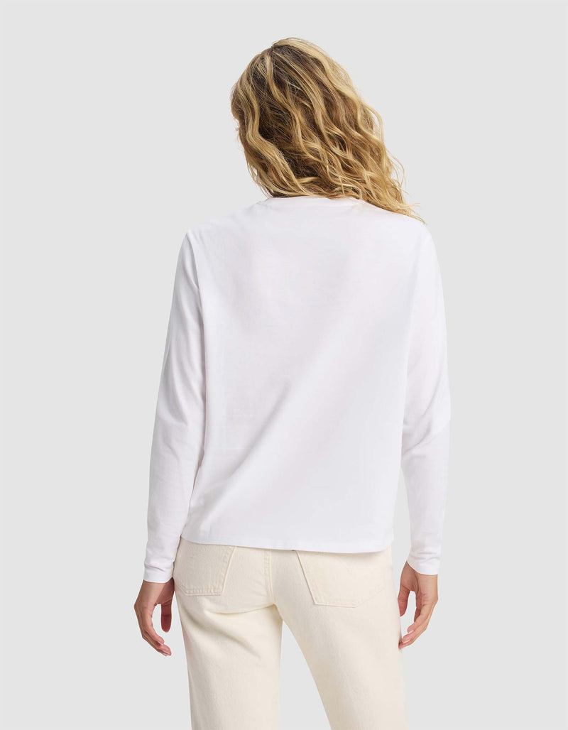 A woman with wavy blonde hair is shown from behind, wearing the Cozy Earth Women's All Day Long Sleeve Tee in white and off-white pants against a plain light gray background.