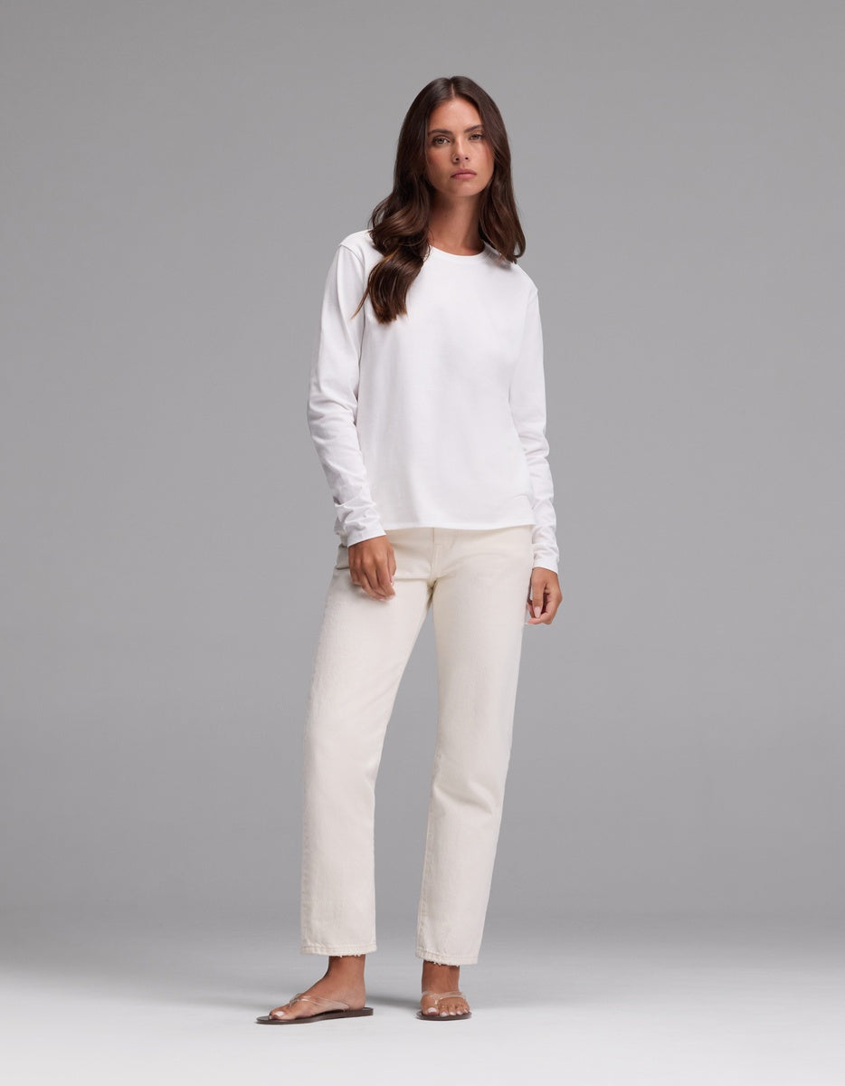 A woman with long brown hair wears the Cozy Earth Women’s All Day Long Sleeve Tee in white, paired with cream-colored pants and sandals, standing against a plain gray background.