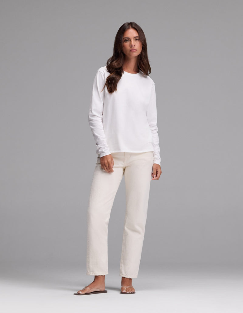 A woman with long brown hair wears the Cozy Earth Women’s All Day Long Sleeve Tee in white, paired with cream-colored pants and sandals, standing against a plain gray background.