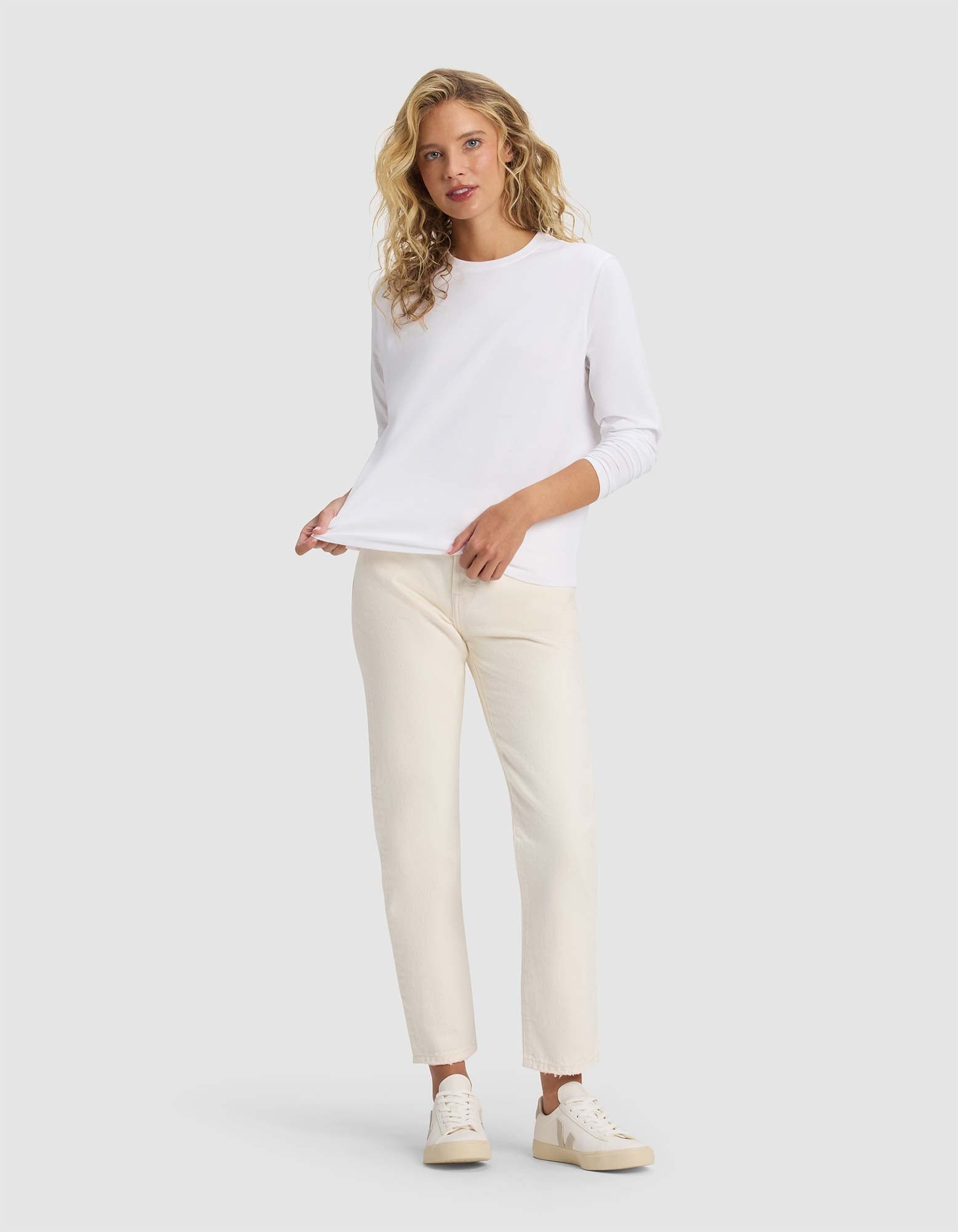 A woman with wavy blonde hair models the Cozy Earth Women's All Day Long Sleeve Tee in white, paired with off-white pants and white sneakers. She stands against a light gray background, looking at the camera with a relaxed expression.