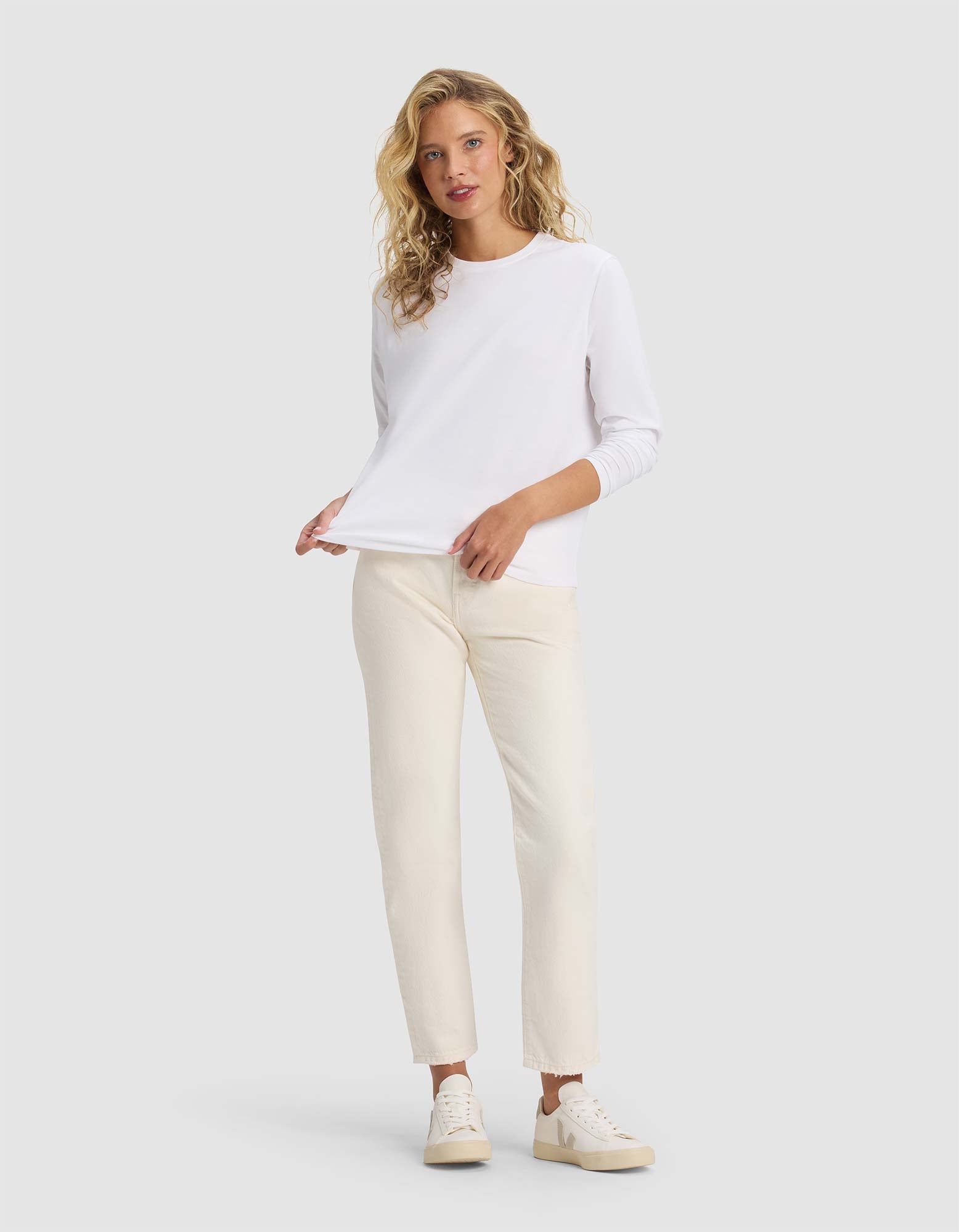 A woman with wavy blonde hair models the Cozy Earth Women's All Day Long Sleeve Tee in white, paired with off-white pants and white sneakers. She stands against a light gray background, looking at the camera with a relaxed expression.