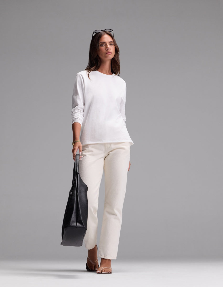 A woman models the Cozy Earth Women’s All Day Long Sleeve Tee in white, paired with matching white pants. She stands holding a large black bag, wearing sandals and sunglasses on her head, against a plain gray background.