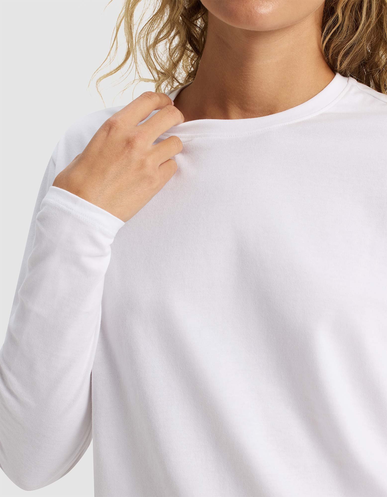 A person with wavy blond hair wears the Cozy Earth Women's All Day Long Sleeve Tee in white, gently holding the collar with one hand. The neutral light gray background and partially out-of-frame face highlight the shirt.|Color:White