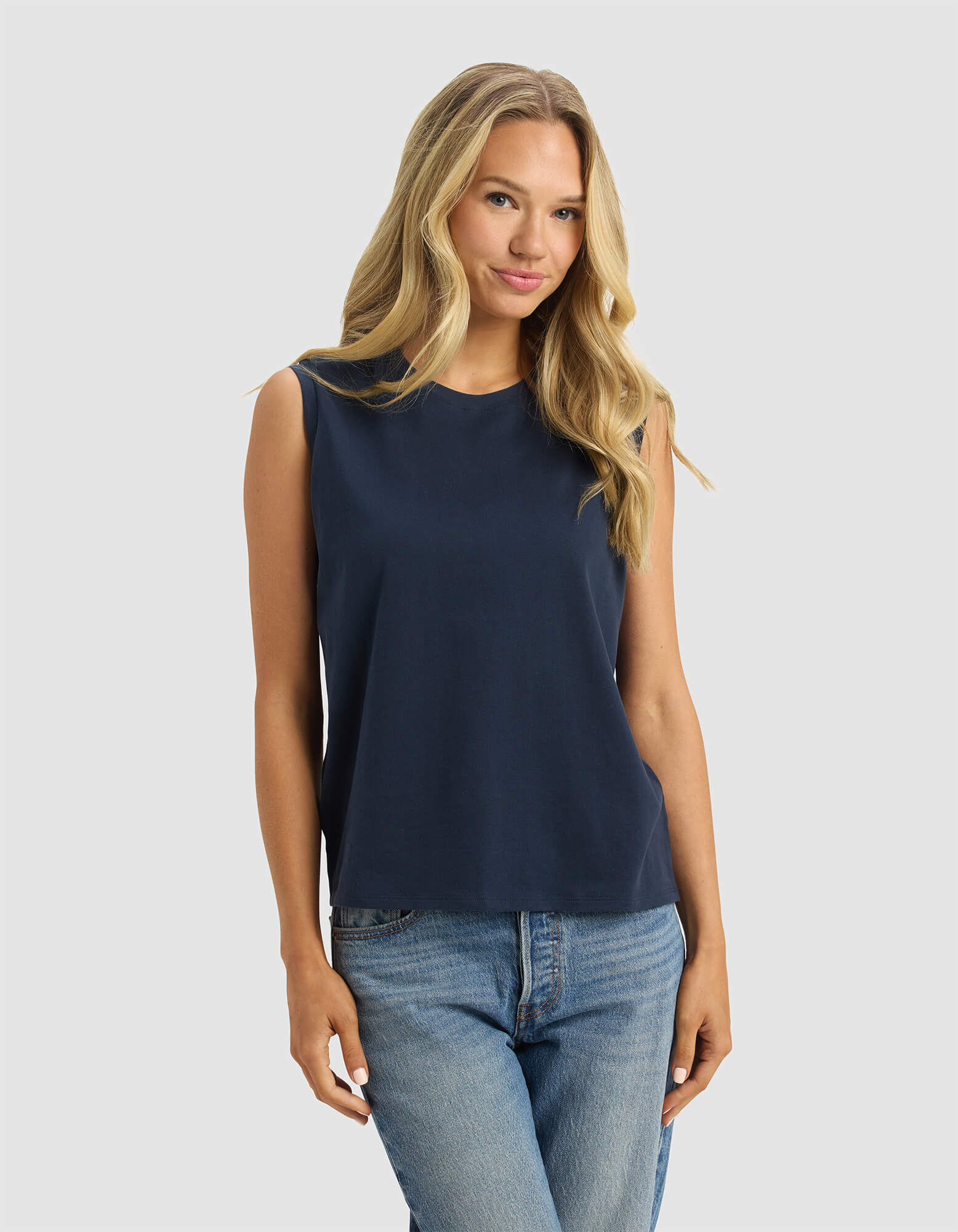 A woman with long blonde hair models the Cozy Earth Women's All Day Sleeveless Tee in navy blue and blue jeans, standing against a plain light gray background and looking forward with a slight smile.