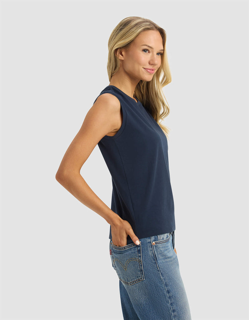 A woman with long blonde hair models the Cozy Earth Women's All Day Sleeveless Tee in navy, paired with blue jeans. She stands sideways with hands in pockets, smiling against a light gray background.