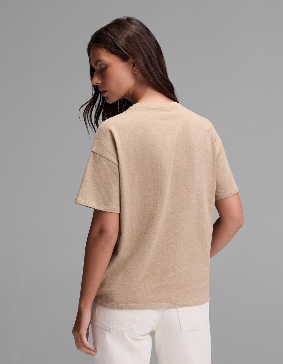 A woman with long brown hair stands facing away, wearing the Cozy Earth Women’s All Day Tee in beige and white pants against a plain gray background.|Color:Beech Heather