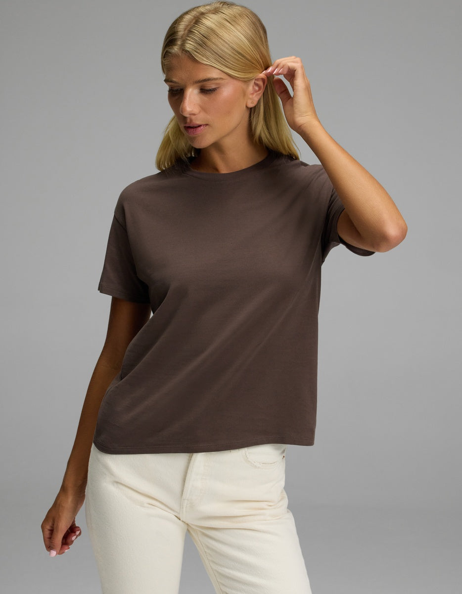 A woman wearing the Cozy Earth Women’s All Day Tee in brown and off-white pants stands against a plain gray background, gently touching her hair and looking down with a neutral expression.