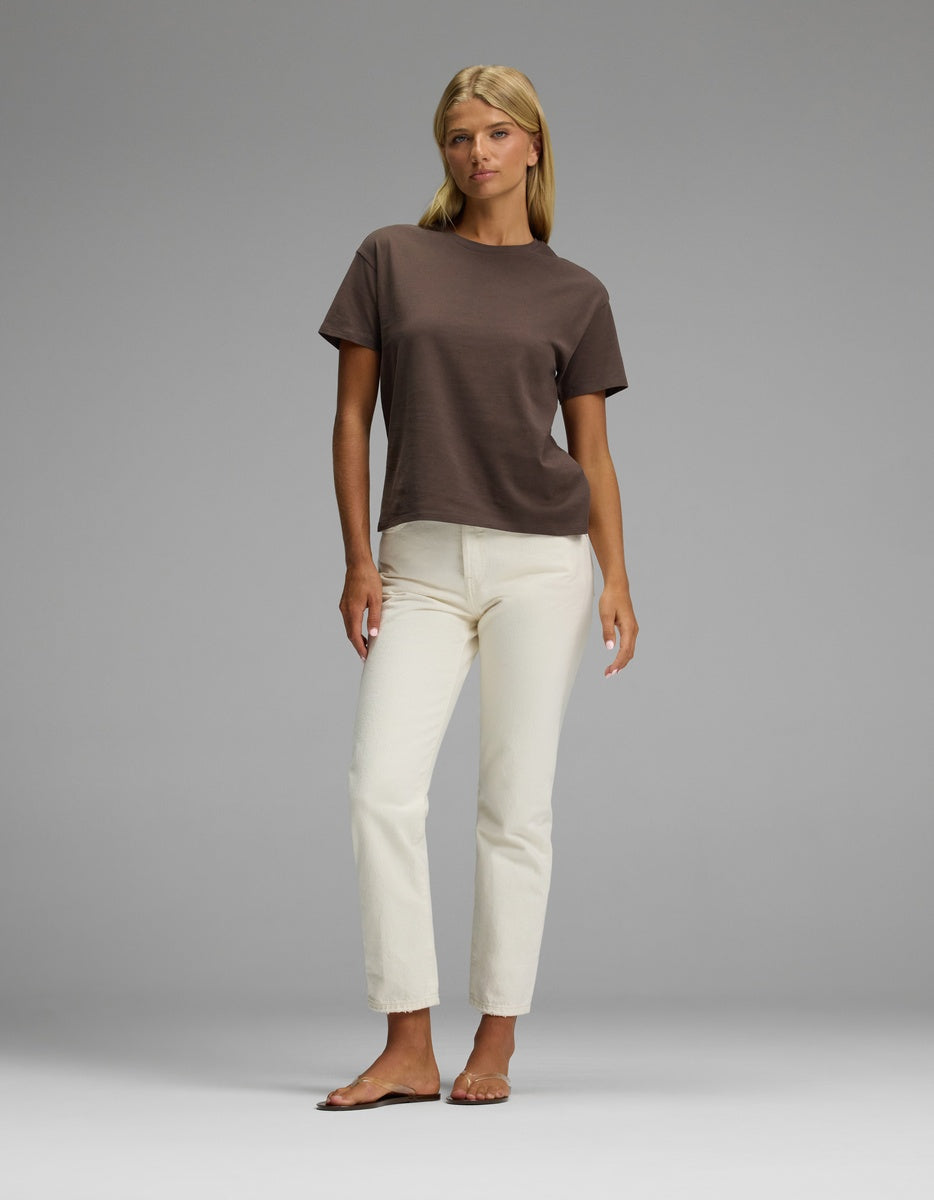 A woman stands barefoot against a gray background, wearing the Cozy Earth Women’s All Day Tee in brown and off-white straight-leg pants, with her arms relaxed at her sides.