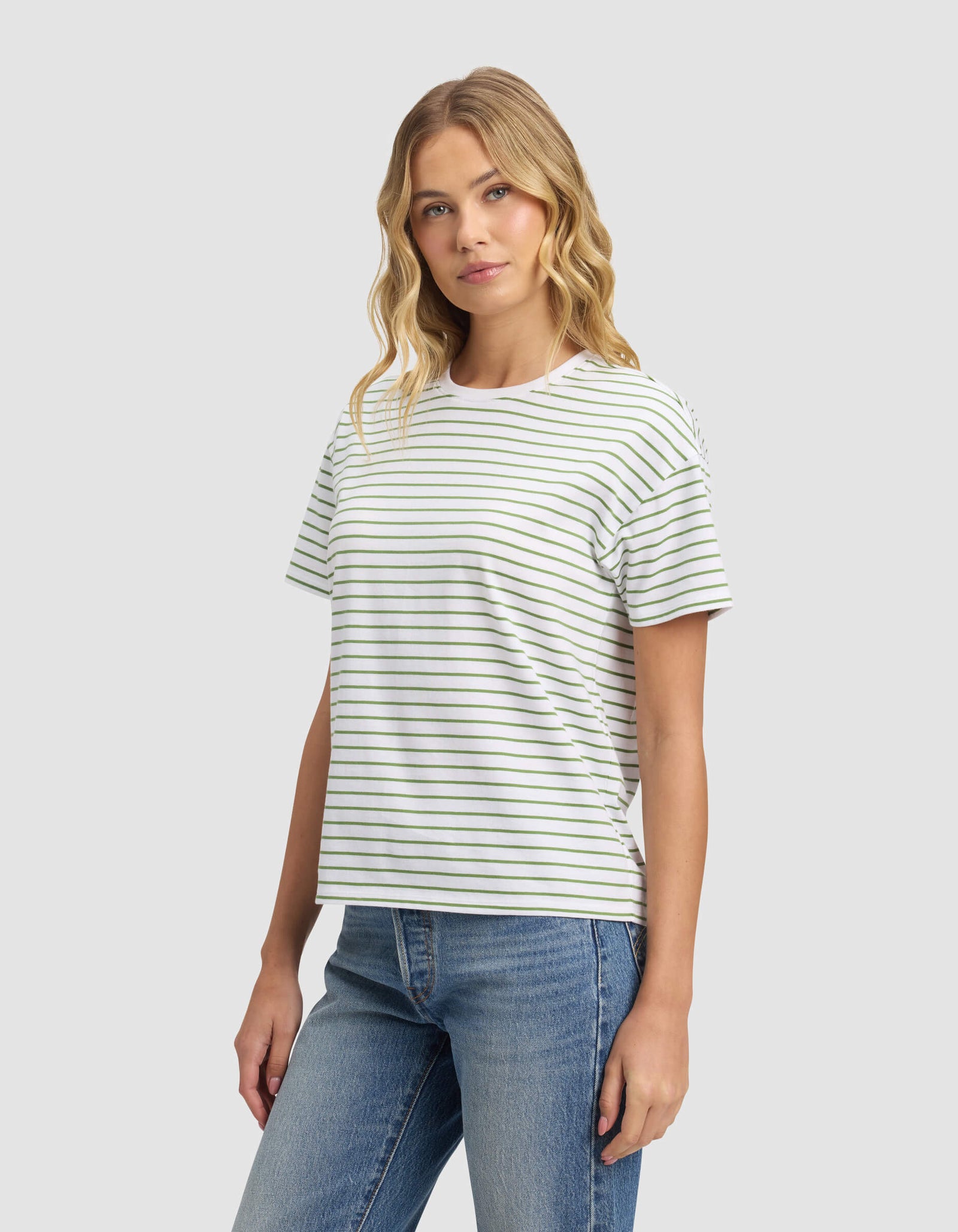 A woman with wavy blonde hair models the Cozy Earth Women's All Day Tee, a white shirt with thin green stripes, paired with blue jeans against a plain light gray background, glancing slightly to the side.