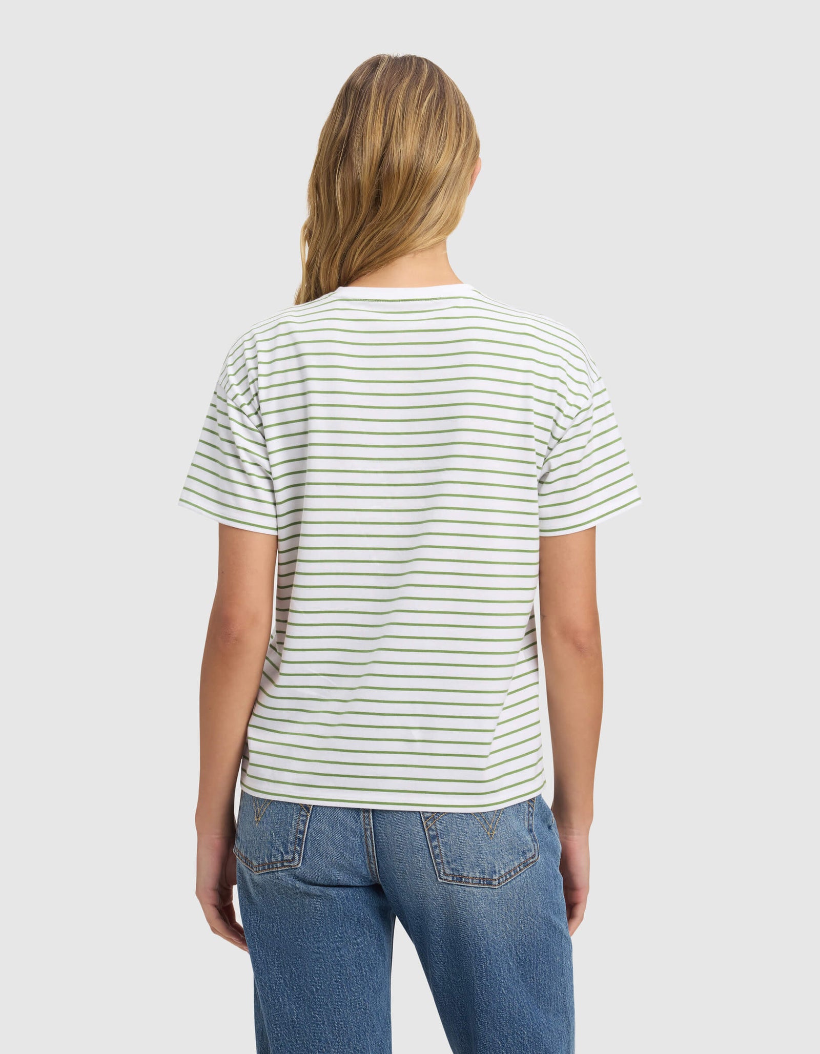 A woman with long, light brown hair faces away wearing the Cozy Earth Women's All Day Tee—white with thin horizontal black stripes—and blue jeans, set against a plain light gray background.