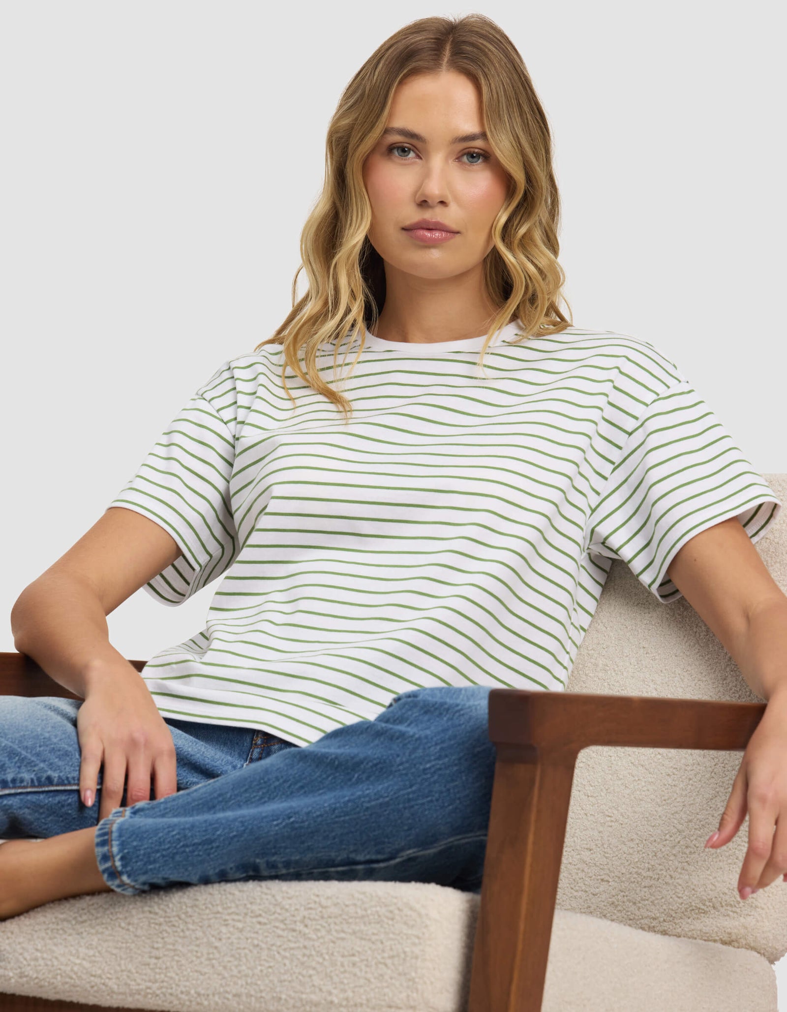 A woman with wavy blonde hair sits on a light-colored armchair, wearing Cozy Earth's Women's All Day Tee and blue jeans, looking directly at the camera with a neutral expression.