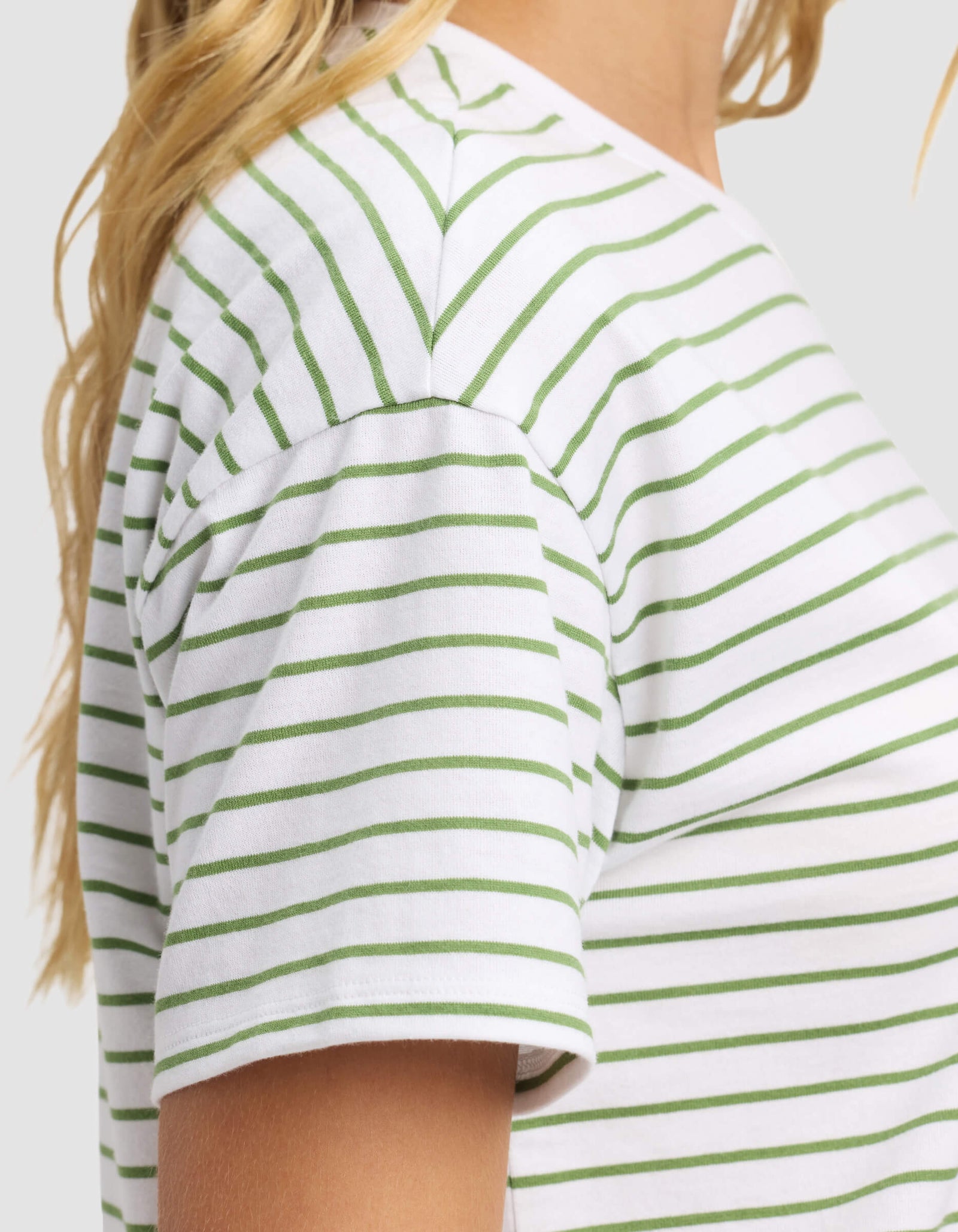 A woman wears the Cozy Earth Women's All Day Tee, a white t-shirt with horizontal green stripes. Only her shoulder, upper arm, and blonde hair are visible against a plain light background.