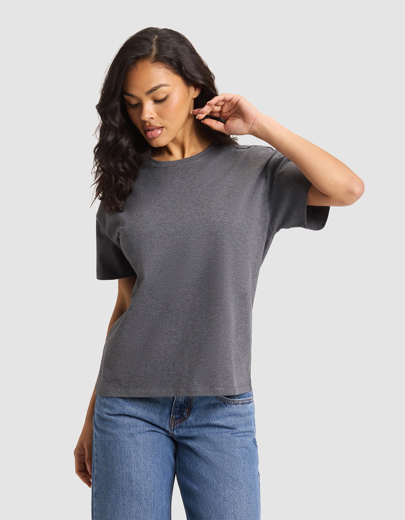 A woman with long, wavy hair wears the Cozy Earth Women's All Day Tee in gray and blue jeans, standing against a light gray background while looking down and touching her ear. 