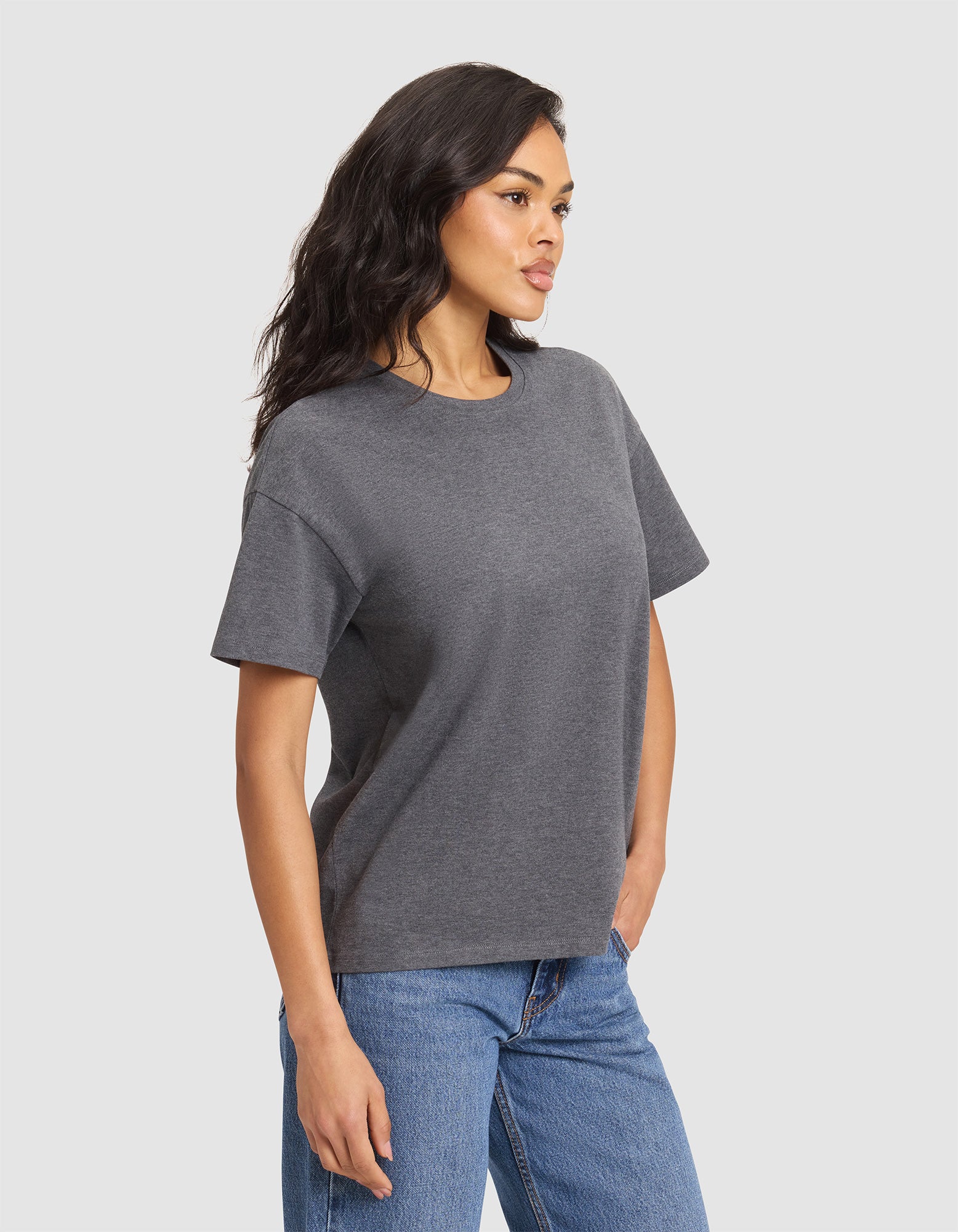 A woman with long dark hair wears the Cozy Earth Women's All Day Tee in dark gray and blue jeans, standing slightly turned left against a light gray background. 