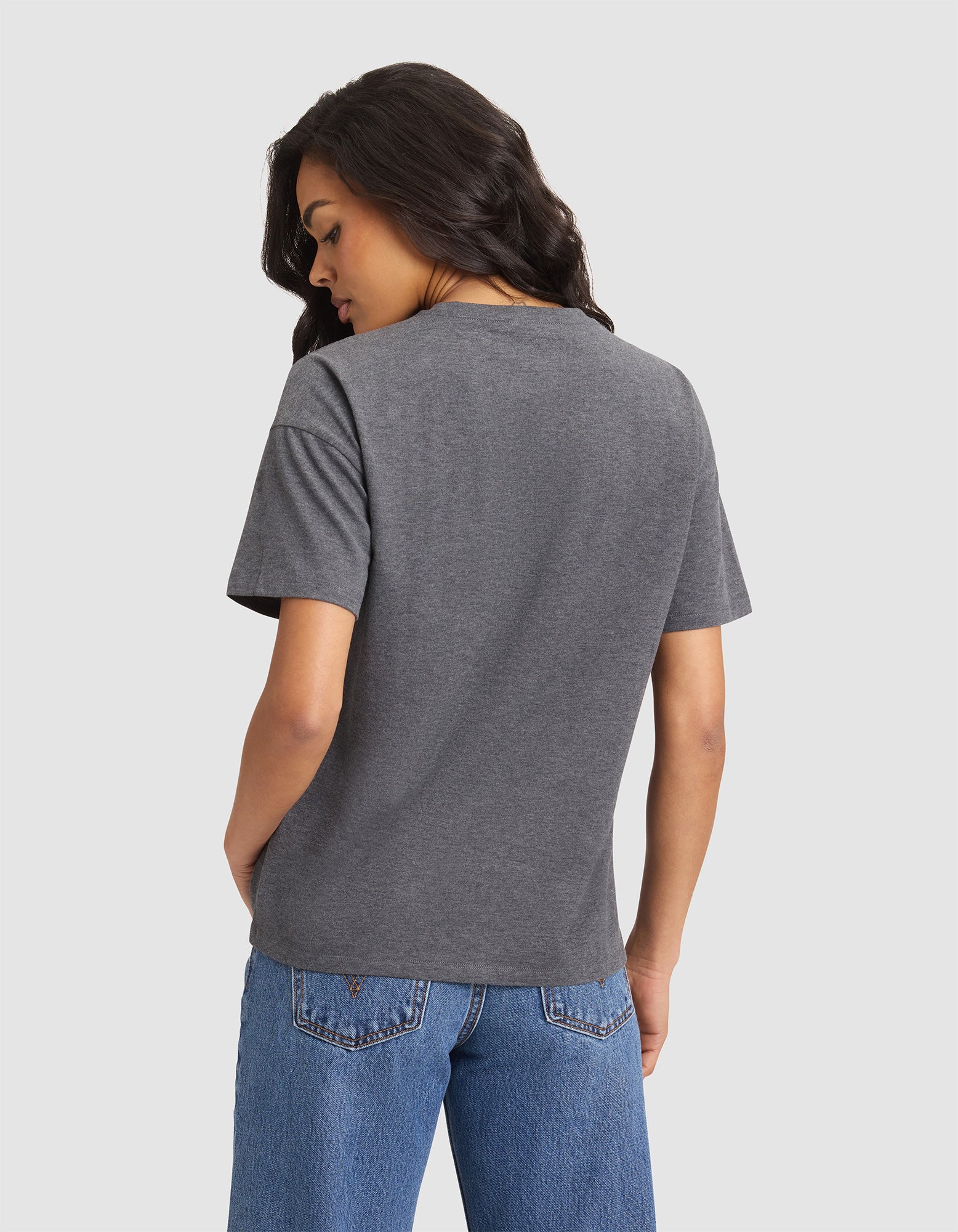 A woman with long dark hair, wearing the Cozy Earth Women's All Day Tee in gray and blue jeans, stands facing away with hands in her pockets against a plain light background. 