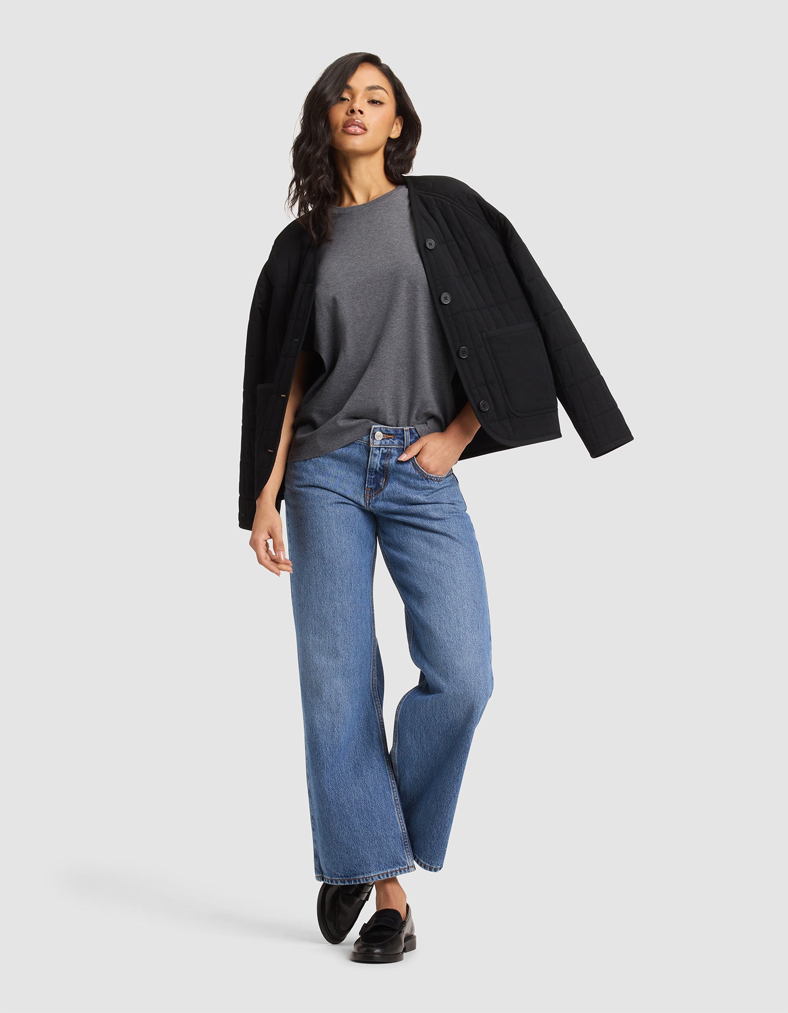 A woman poses in a Cozy Earth Women's All Day Tee, paired with a black quilted jacket, blue straight-leg jeans, and black loafers. She has long, wavy dark hair and stands against a neutral background. 