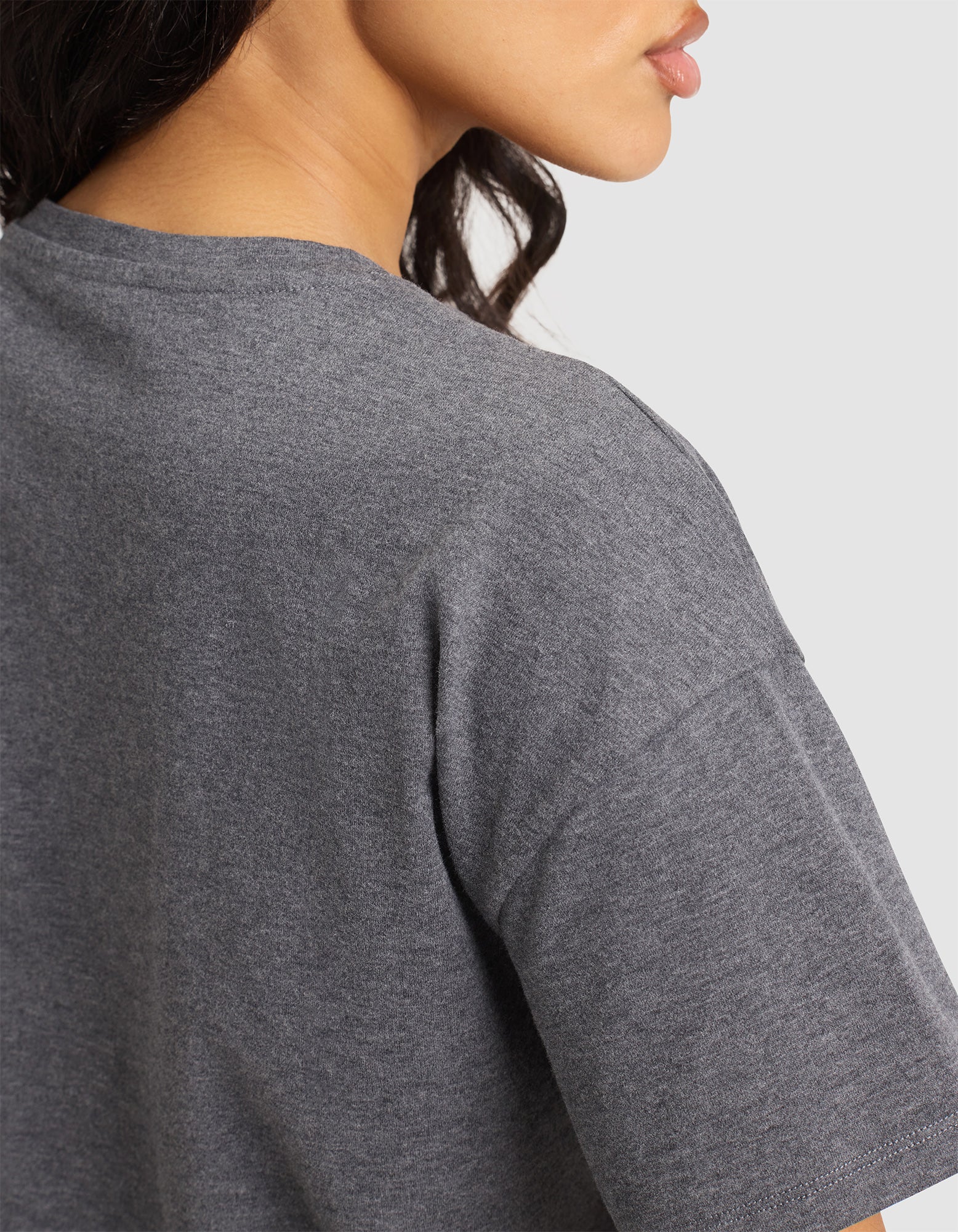 A close-up shows a woman with long, wavy dark hair wearing the Cozy Earth Women's All Day Tee in heather gray, back and left sleeve visible. Her face is partly in profile against a plain light gray background. 