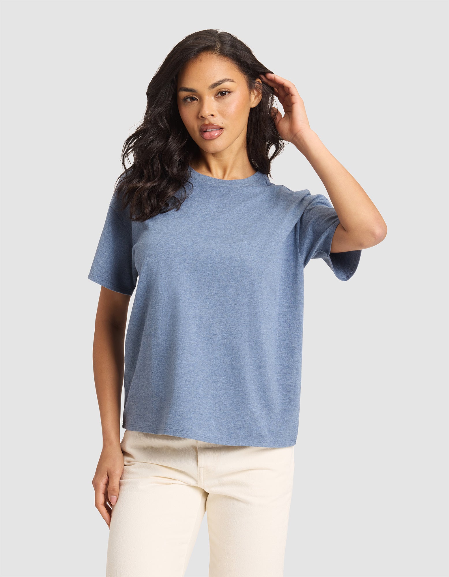 A woman with long dark hair wears the Cozy Earth Women's All Day Tee in light blue and off-white pants, standing against a plain light gray background. She faces the camera, touching her hair with one hand.  