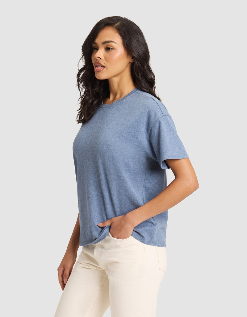 A woman with wavy dark hair models the Cozy Earth Women's All Day Tee, a loose-fitting blue short-sleeve t-shirt, paired with light-colored pants, standing with one hand in her pocket against a plain light gray background.