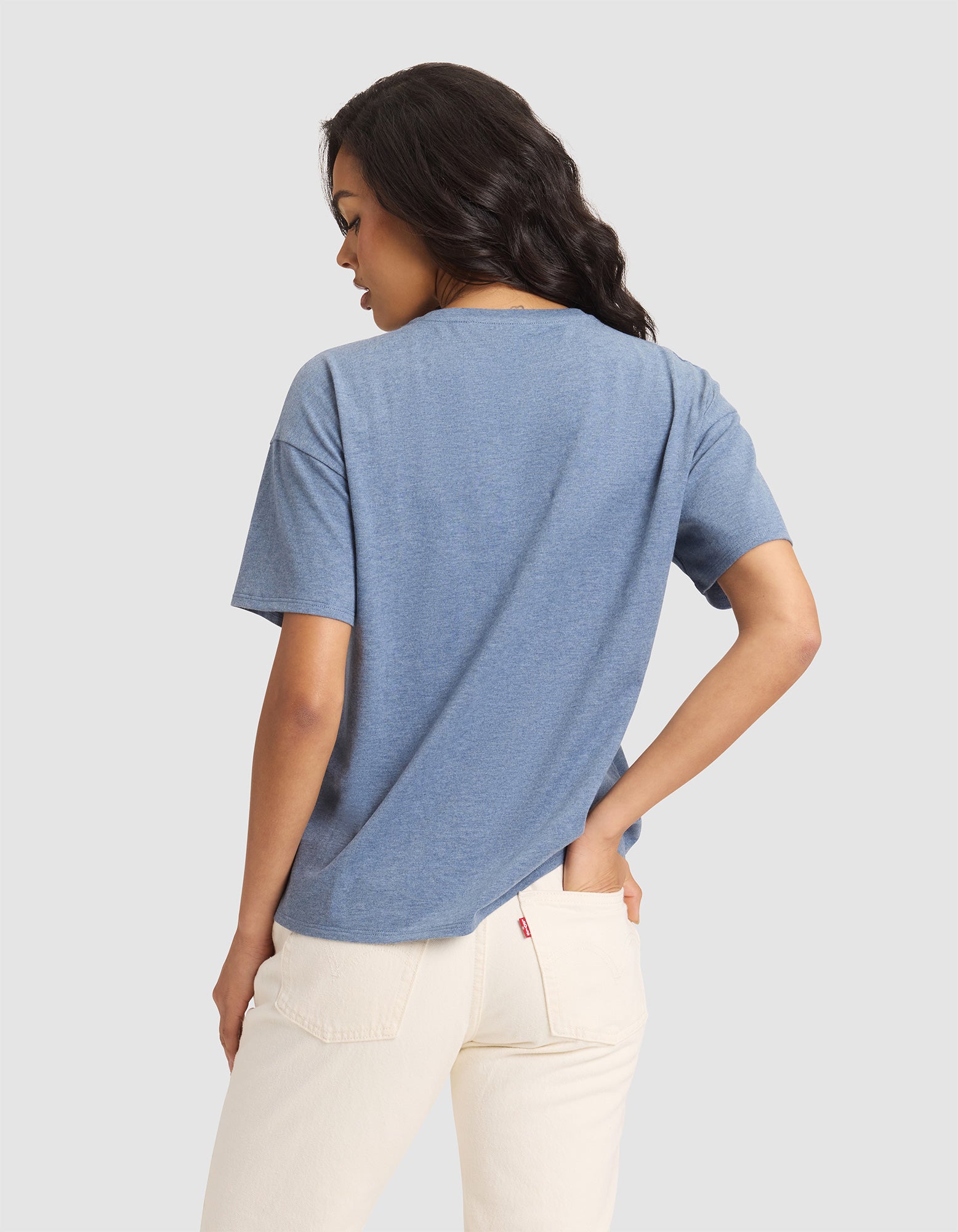 A woman with long, dark hair faces away from the camera wearing the Cozy Earth Women's All Day Tee in blue and light-colored jeans, set against a plain light gray background. 
