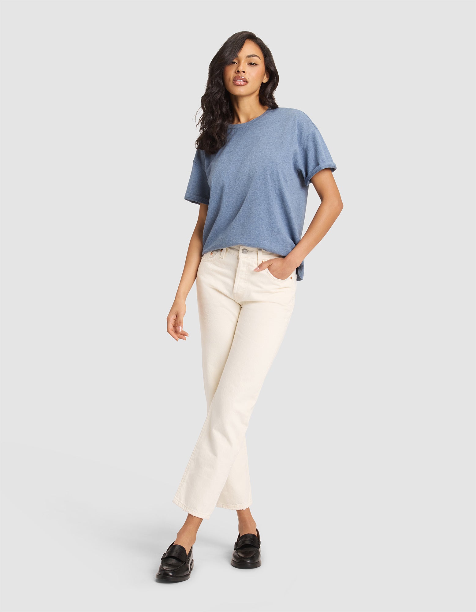 Against a plain backdrop, a woman with long, wavy dark hair wears the Cozy Earth Women's All Day Tee in blue, off-white straight-leg jeans, and black loafers, casually posing with one hand in her pocket. 
