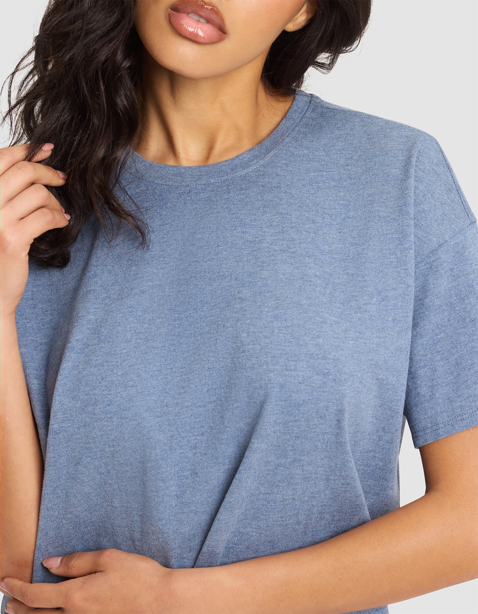 A woman with wavy dark hair models the Cozy Earth Women's All Day Tee, a light blue short-sleeve crew neck, cropped from the shoulders down against a plain background. One hand touches her hair, the other rests at her side. 