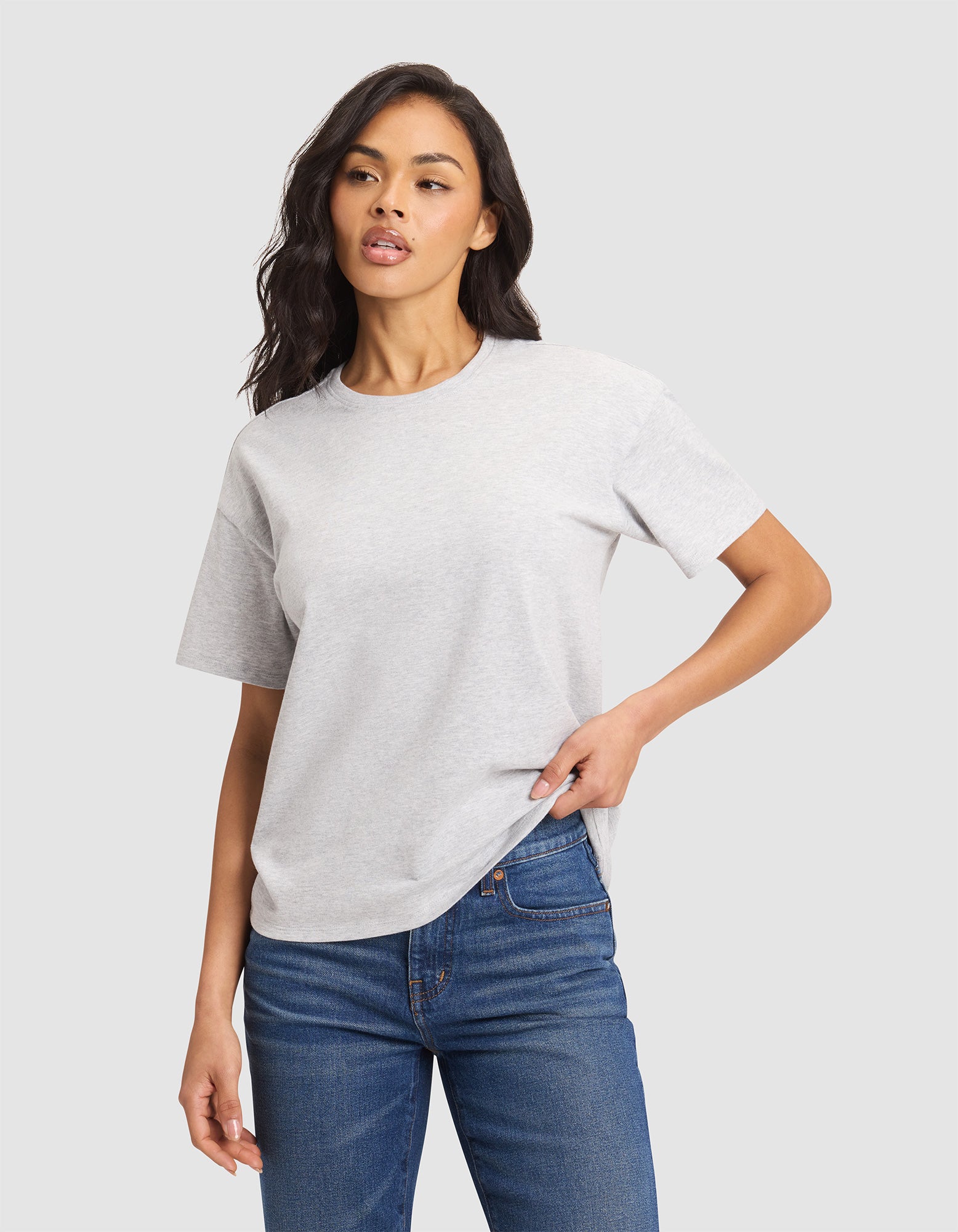 A woman wears the Cozy Earth Women's All Day Tee in light gray with blue jeans, standing against a plain light background, one hand in her pocket and the other relaxed by her side. |Color:French Dove Heather