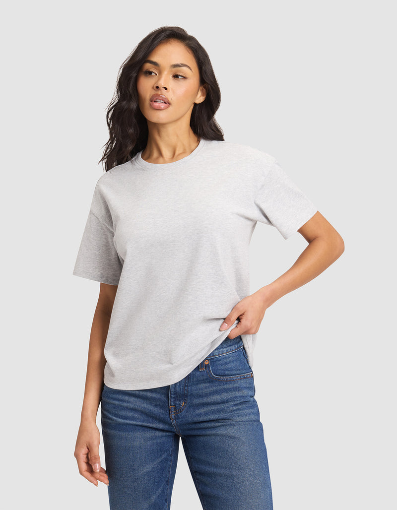 A woman wears the Cozy Earth Women's All Day Tee in light gray with blue jeans, standing against a plain light background, one hand in her pocket and the other relaxed by her side.