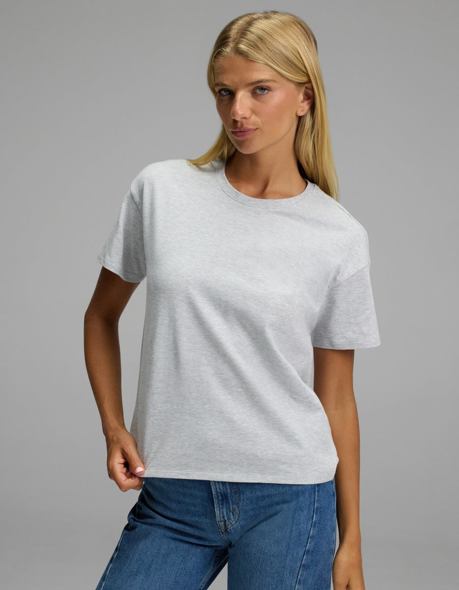 A woman with long blonde hair wearing the Cozy Earth Women’s All Day Tee in light gray and blue jeans stands against a plain gray background, looking confidently at the camera.|Color:Dove Grey Heather