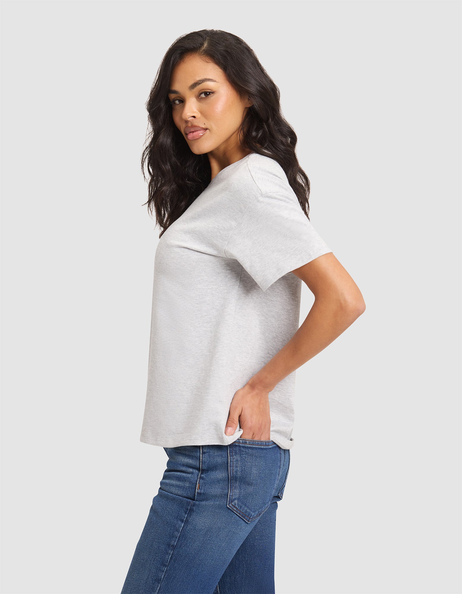 A woman with long dark hair wears the Cozy Earth Women's All Day Tee in light gray and blue jeans, standing in profile with one hand in her back pocket against a plain light background. 