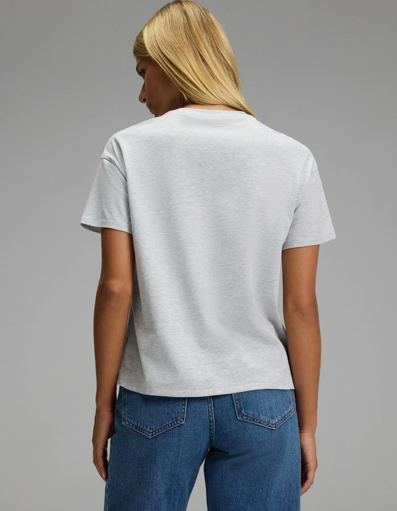 A person with straight blonde hair wears the Cozy Earth Women’s All Day Tee in light gray, paired with blue jeans, standing with their back to the camera against a plain gray background.