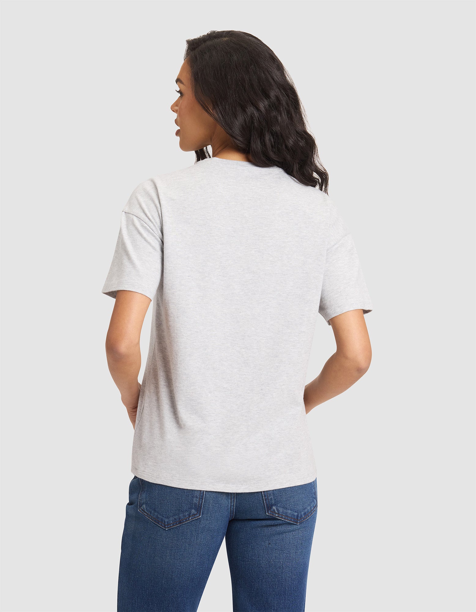 A woman stands facing away with her hands on her hips, wearing the Cozy Earth Women's All Day Tee in light gray and blue jeans, against a plain light gray background. 