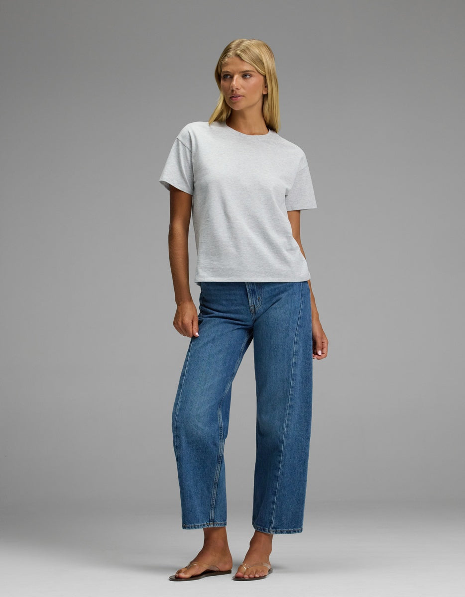 A woman stands barefoot against a plain gray background, wearing the Cozy Earth Women’s All Day Tee in light gray and blue jeans. She has straight blonde hair and gazes slightly to her left.