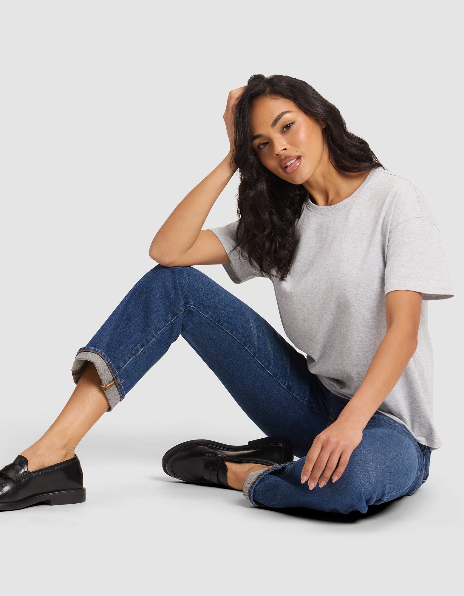 A woman sits on the floor with one knee up, wearing Cozy Earth’s Women's All Day Tee in light gray, blue rolled-up jeans, and black loafers. Her left hand rests on her head and she looks relaxed. 