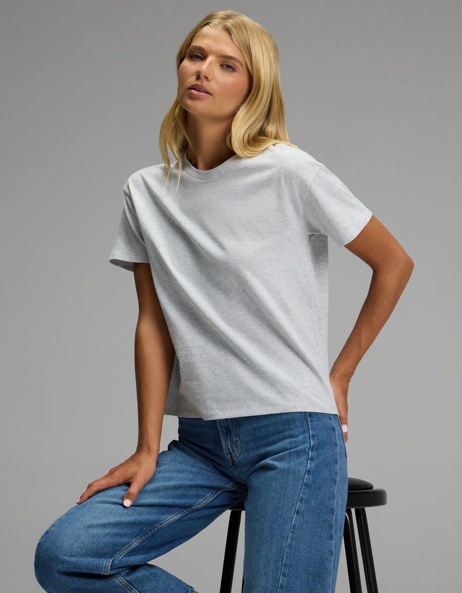 A woman with long blonde hair sits on a stool against a plain gray background, wearing the Cozy Earth Women’s All Day Tee in light gray and blue jeans.