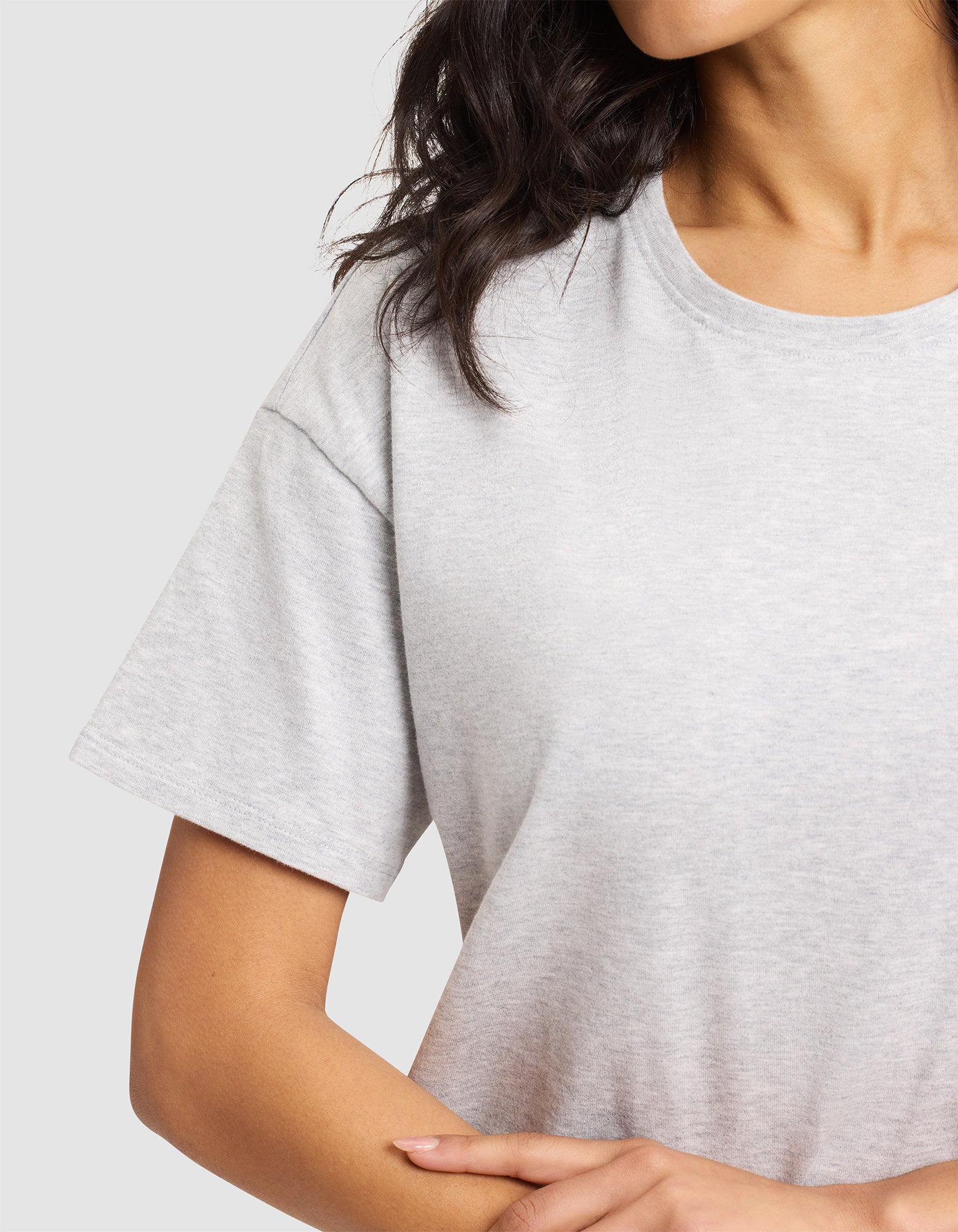 A woman wears the Cozy Earth Women's All Day Tee in light gray with short sleeves, standing with one arm bent and her hand on her side. Her wavy dark hair falls over her shoulder against a plain background. Face is partially out of frame. 