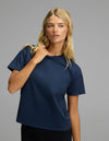 A woman with long blonde hair wears the Cozy Earth Women’s All Day Tee in navy blue and black pants, standing against a plain gray background. She is looking slightly to the side, with one hand touching her collar.|Color:Eclipse