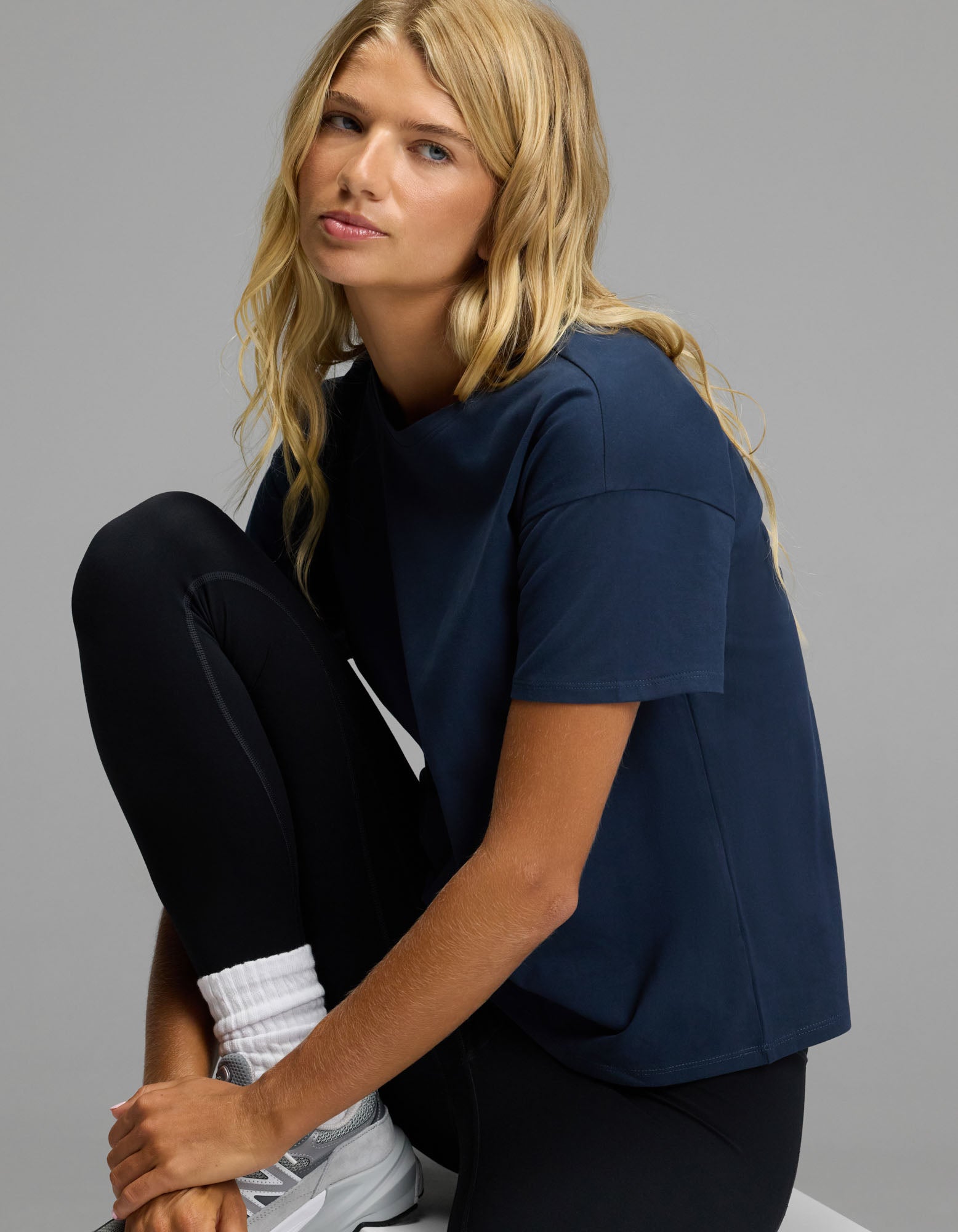 A person with long blonde hair wears the Cozy Earth Women’s All Day Tee in navy, paired with black leggings and white sneakers, sitting with one knee up against a plain gray background.|Color:Eclipse