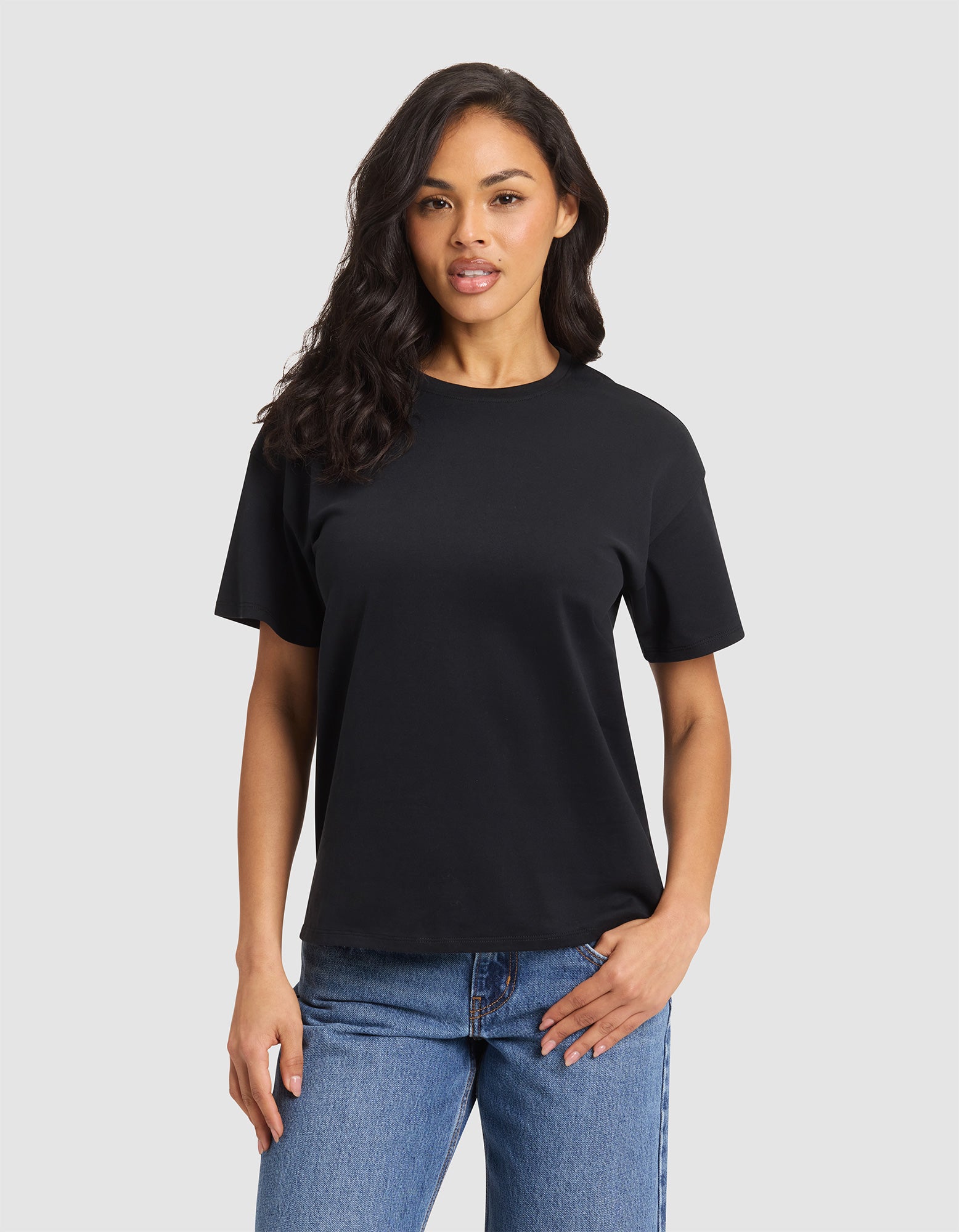 A woman with long dark hair wears the Cozy Earth Women's All Day Tee in black and blue jeans, standing against a light gray background. She faces forward with one hand in her pocket and a neutral expression. 