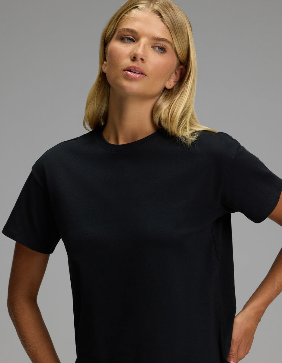 A woman with long blonde hair wears the Women’s All Day Tee by Cozy Earth in black and gazes slightly to the side against a plain gray background.