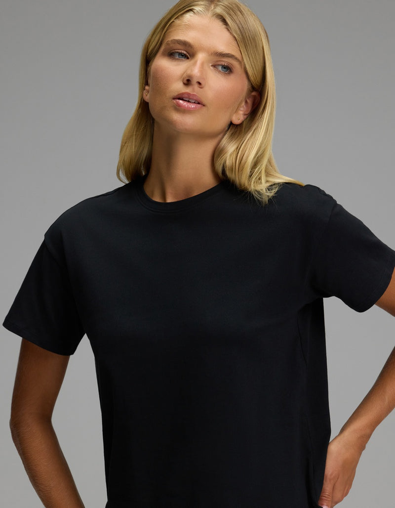A woman with long blonde hair wears the Women’s All Day Tee by Cozy Earth in black and gazes slightly to the side against a plain gray background.