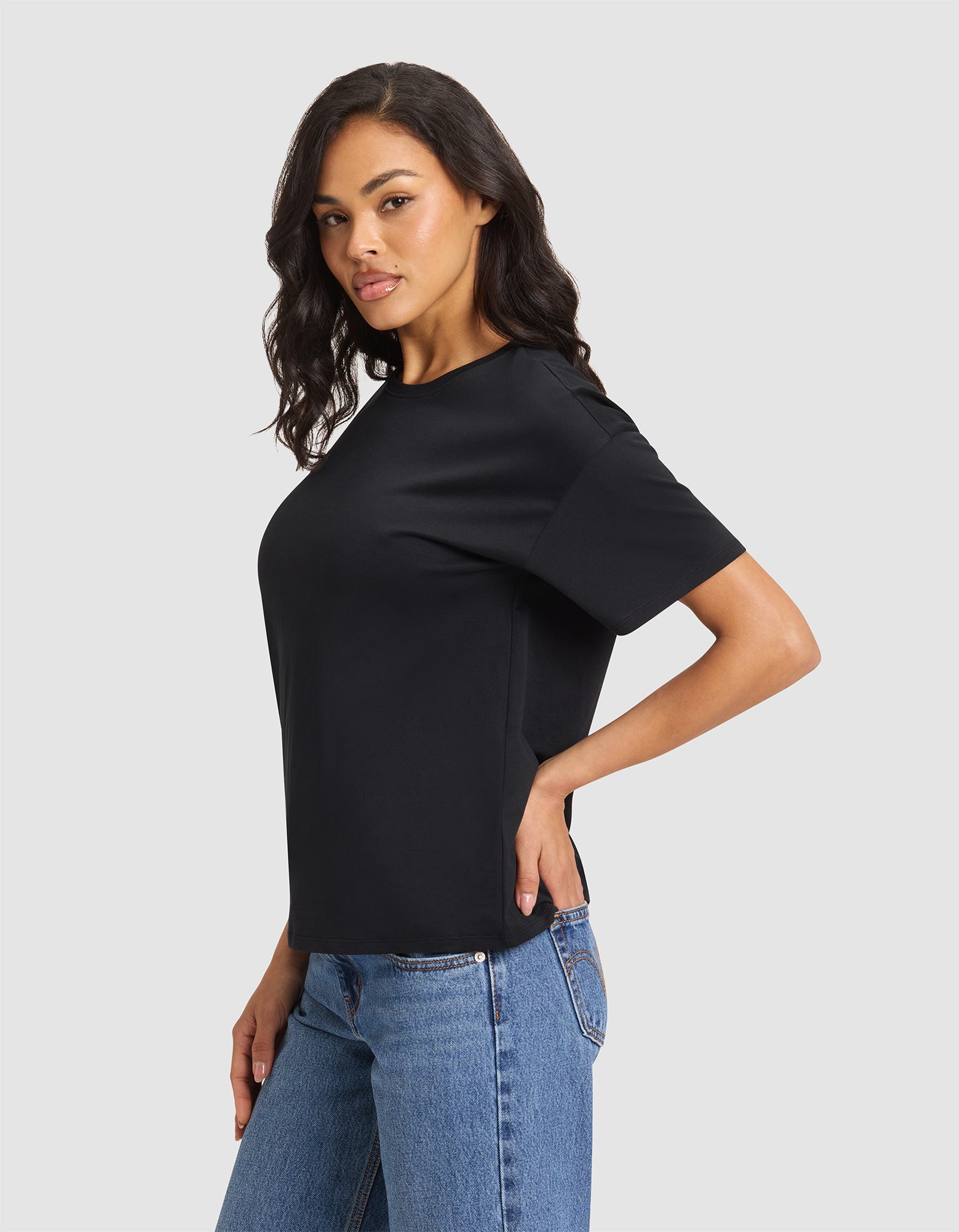 A woman with wavy dark hair models Cozy Earth's Women's All Day Tee, a plain black short-sleeve shirt, paired with blue jeans. She stands sideways with one hand on her hip against a light gray background.  