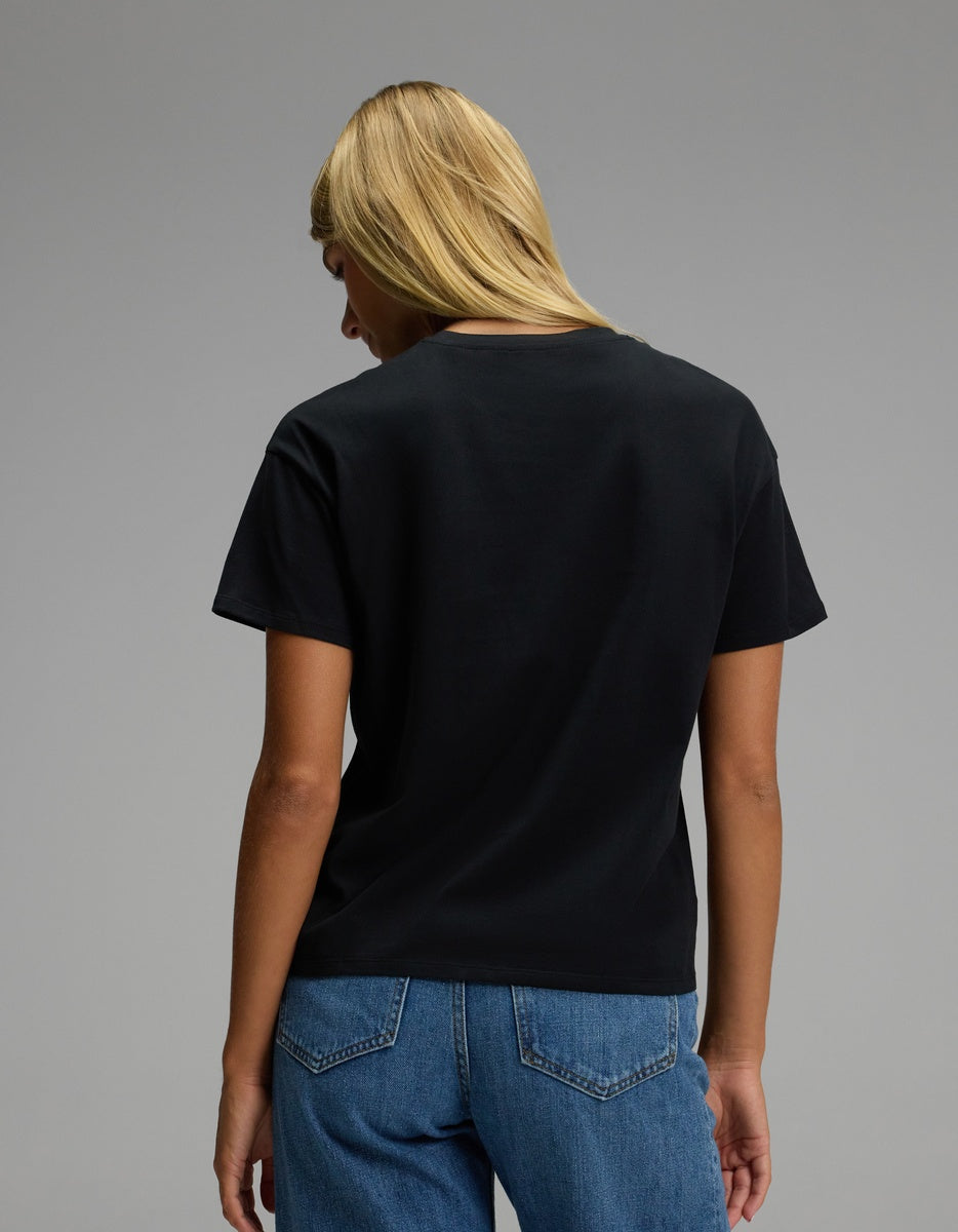 A woman with long blond hair, wearing the Cozy Earth Women’s All Day Tee in black and blue jeans, stands facing away from the camera against a plain gray background.