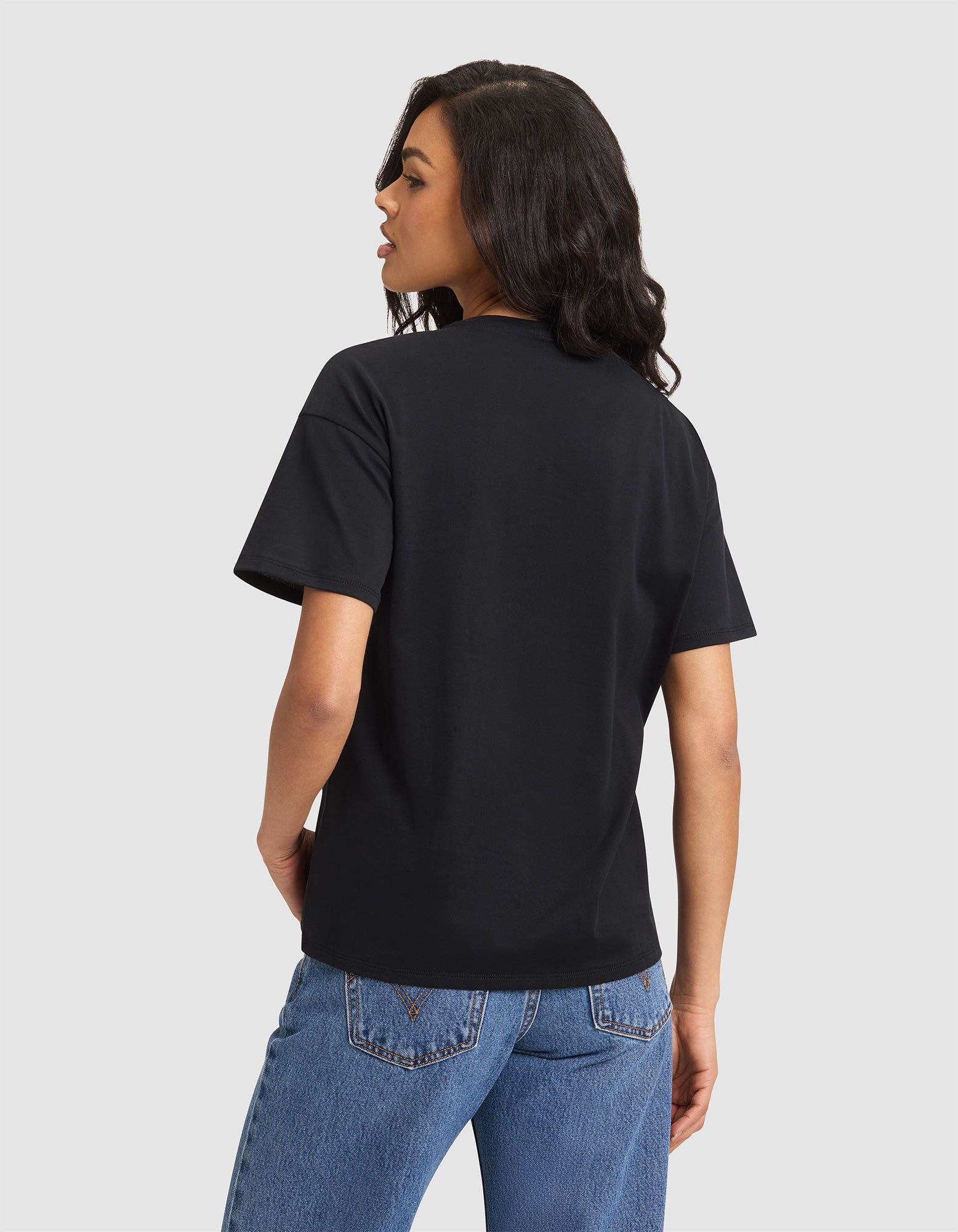 A woman with long dark hair, wearing the Cozy Earth Women's All Day Tee in black and blue jeans, stands with her back to the camera against a light gray background. 
