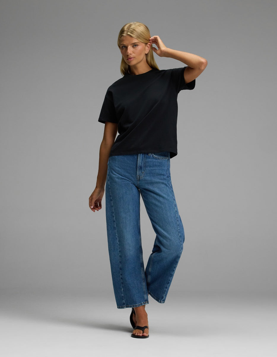 A woman poses in the Cozy Earth Women’s All Day Tee, styled with blue wide-leg jeans and black heeled sandals, against a plain gray background.