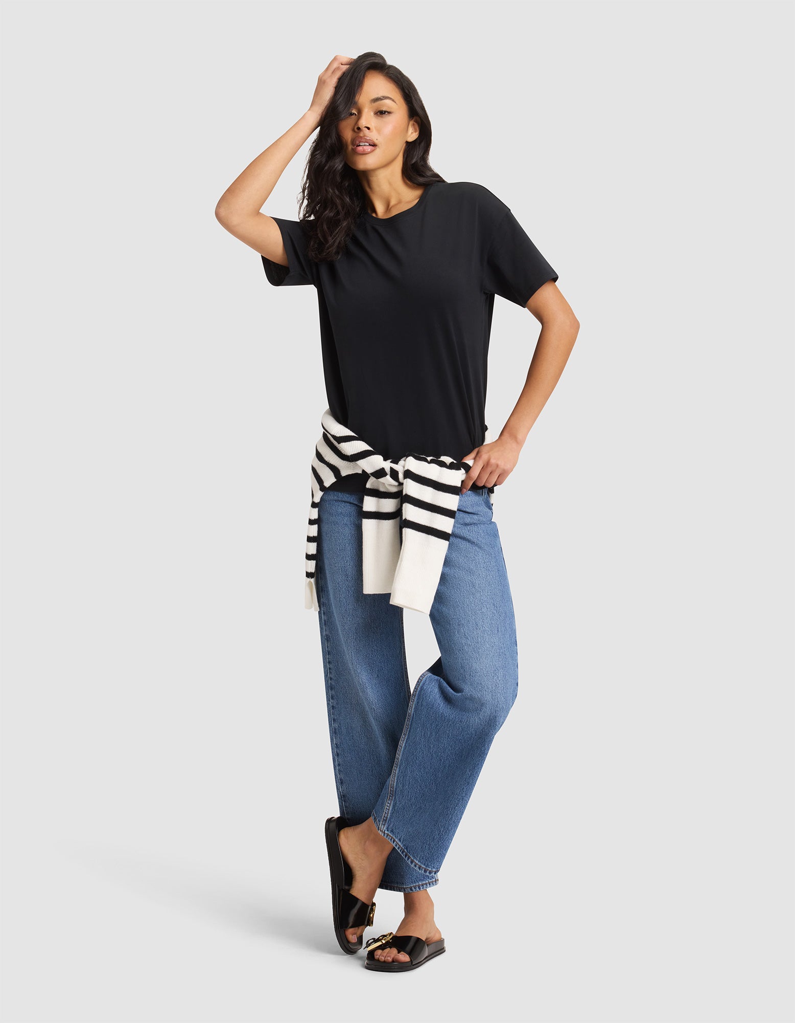 A woman wears the Cozy Earth Women's All Day Tee in black with blue jeans, black sandals, and a striped sweater tied at her waist. She stands against a plain background, touching her hair and smiling softly. 