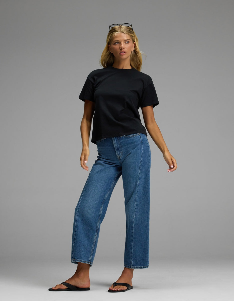 A woman models the Cozy Earth Women’s All Day Tee in black, paired with blue jeans and black sandals against a simple gray background. With blonde hair and sunglasses resting on her head, she gazes slightly to the side with a neutral expression.