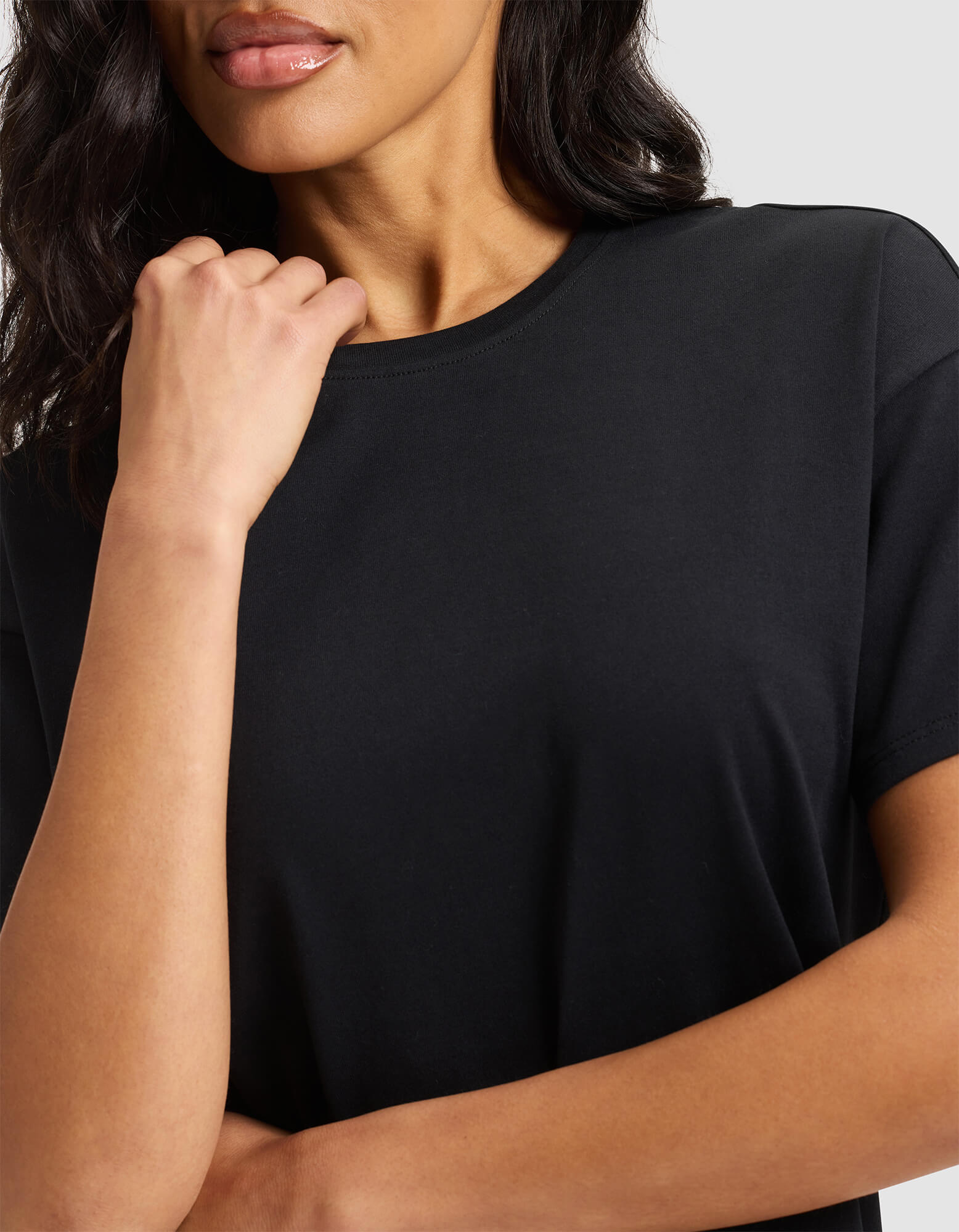 A woman with dark wavy hair poses in the Cozy Earth Women's All Day Tee, a plain black t-shirt. Her left hand rests near her chin, her face is partially visible, and she stands against a neutral background. 