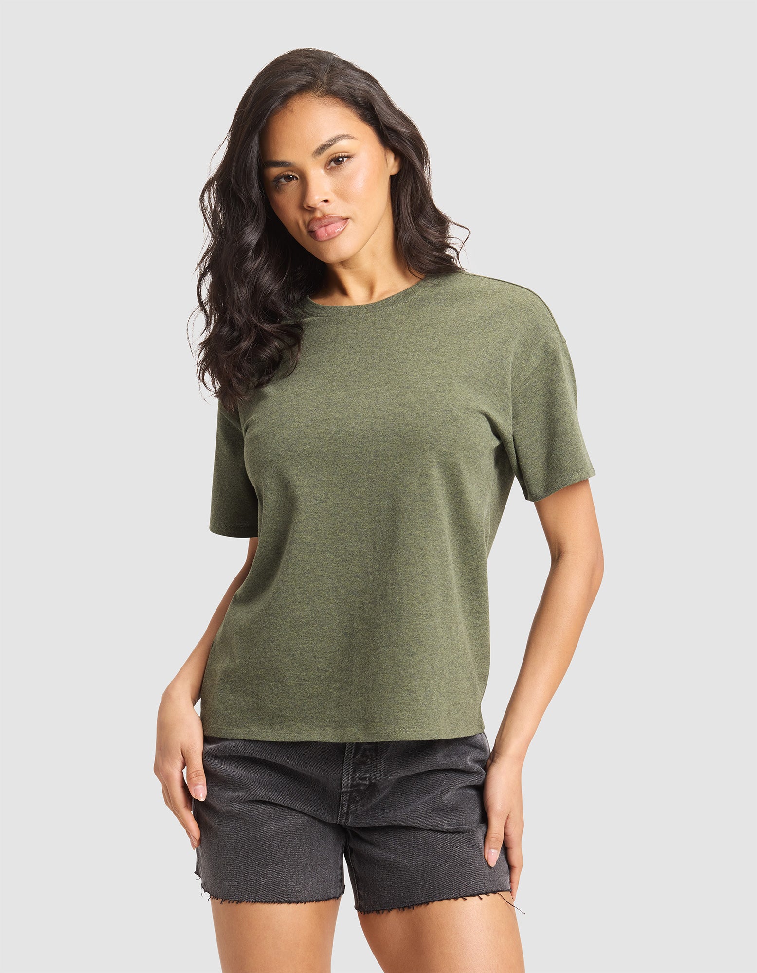 A woman with long dark hair wears the Cozy Earth Women's All Day Tee in green and black denim shorts, standing against a plain light gray background facing forward with one hand on her hip. 