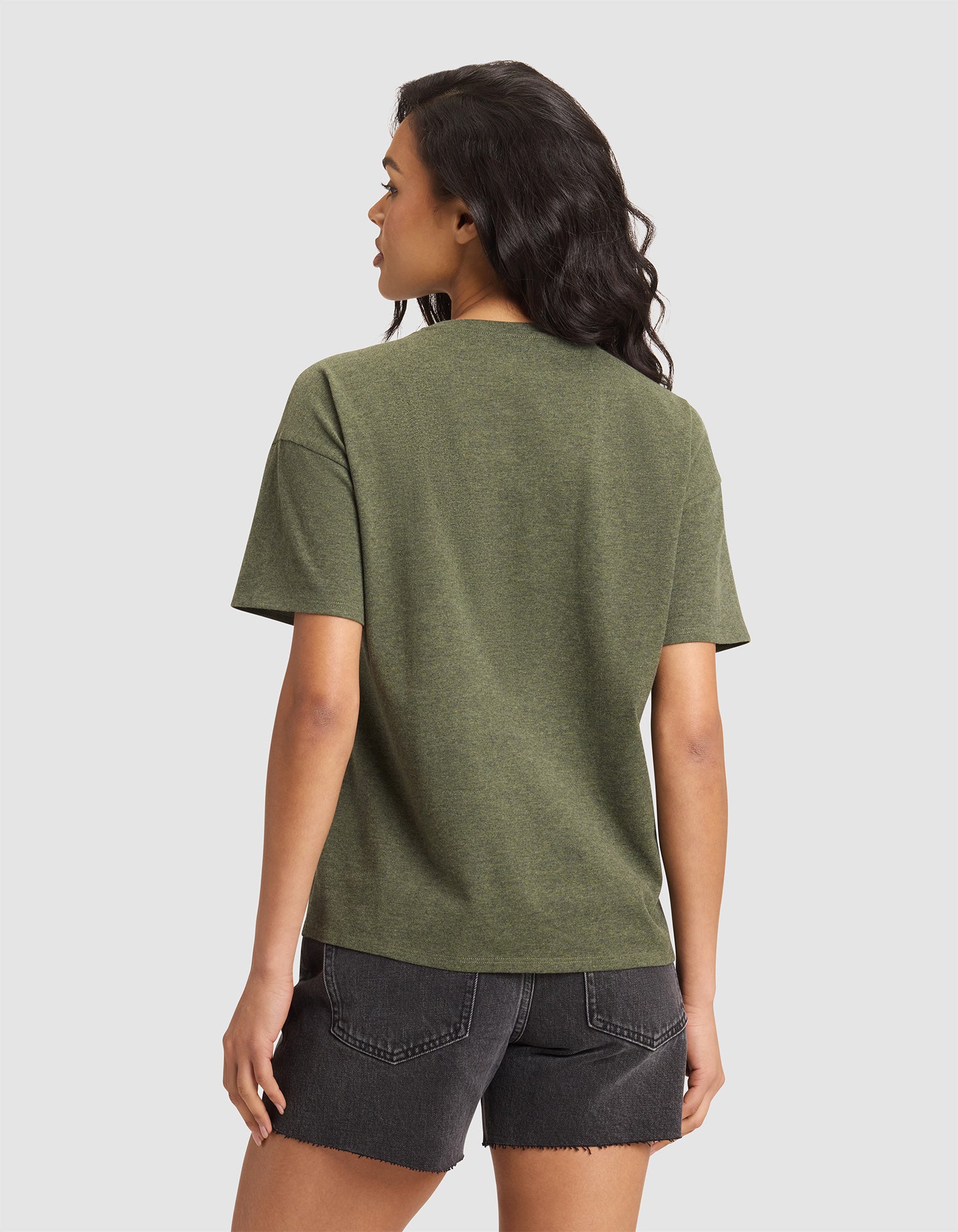 A woman with wavy dark hair stands facing away, wearing the Cozy Earth Women's All Day Tee in green and black denim shorts, set against a plain light gray background. 